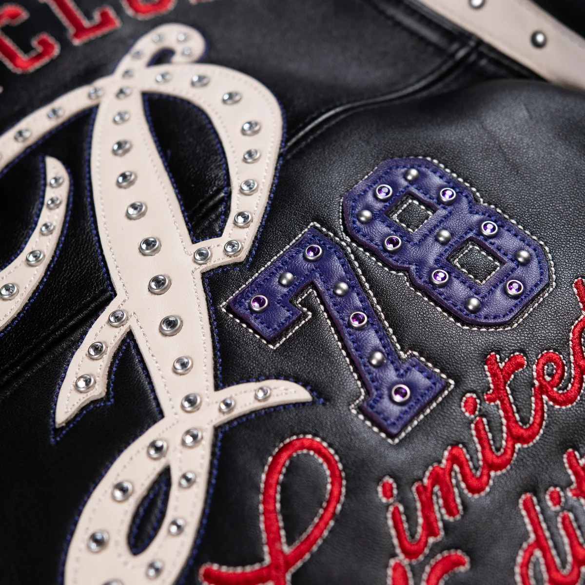 Close view of cream script and purple 78 applique on a black leather jacket, showing silver-tone studs and red embroidered lettering.