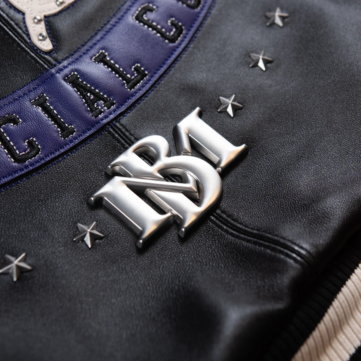 Detail of silver-tone monogram and star embellishments on a black leather jacket, below a purple Special Cut patch with stitched edges.