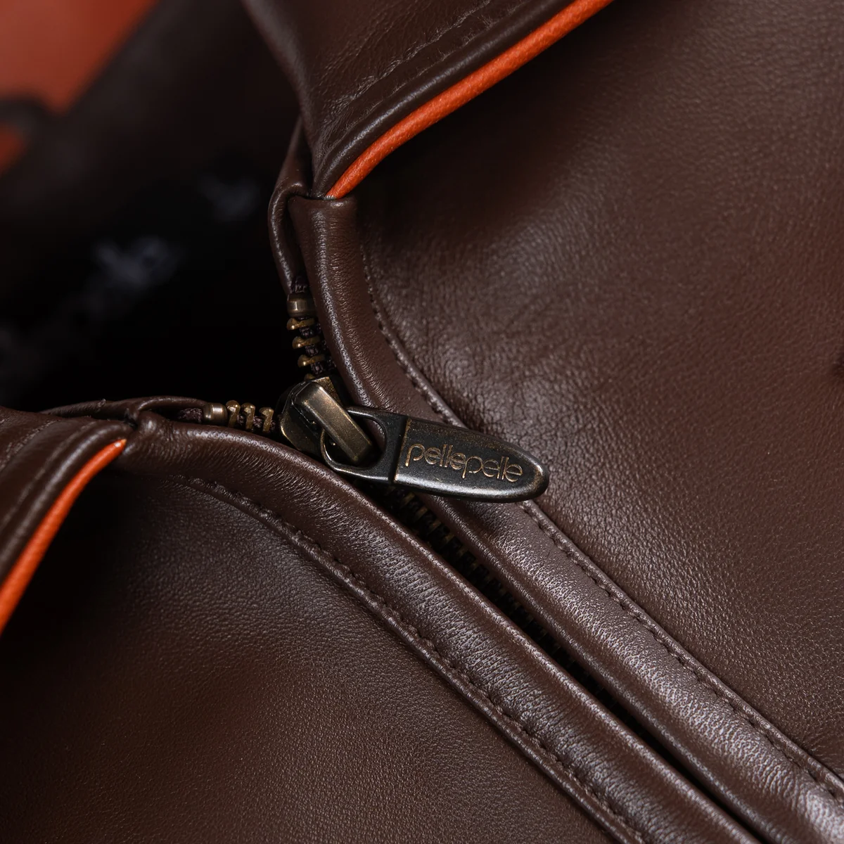 Close view of the brown leather racing jacket collar and front zipper, showing orange piping, visible stitching and branded zipper pull.