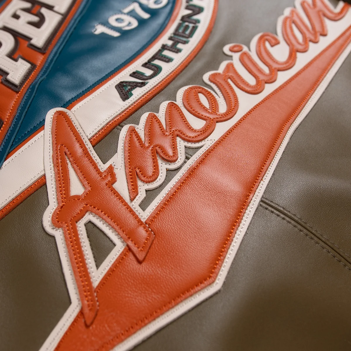 Close view of orange American script applique on the olive racing jacket back, with cream edging, visible stitching and layered panels.