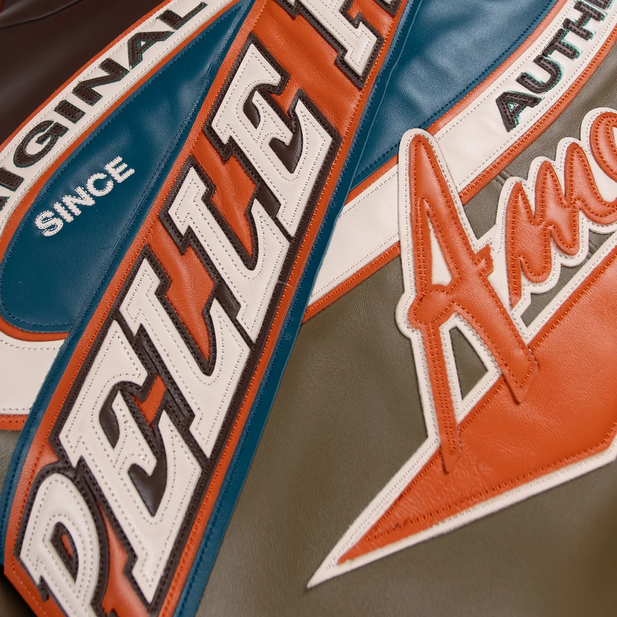 Detailed view of layered Pelle Pelle lettering on the racing sport jacket, showing orange, teal and cream panels with stitched edges.