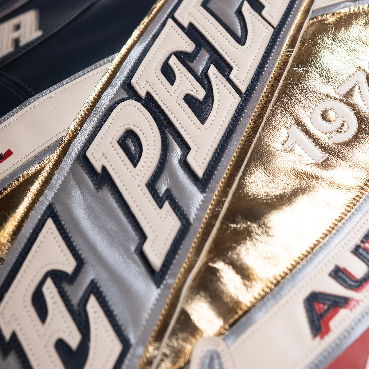 Close view of layered cream lettering across the jacket's metallic silver and gold back patch, showing stitched edges and panel texture.