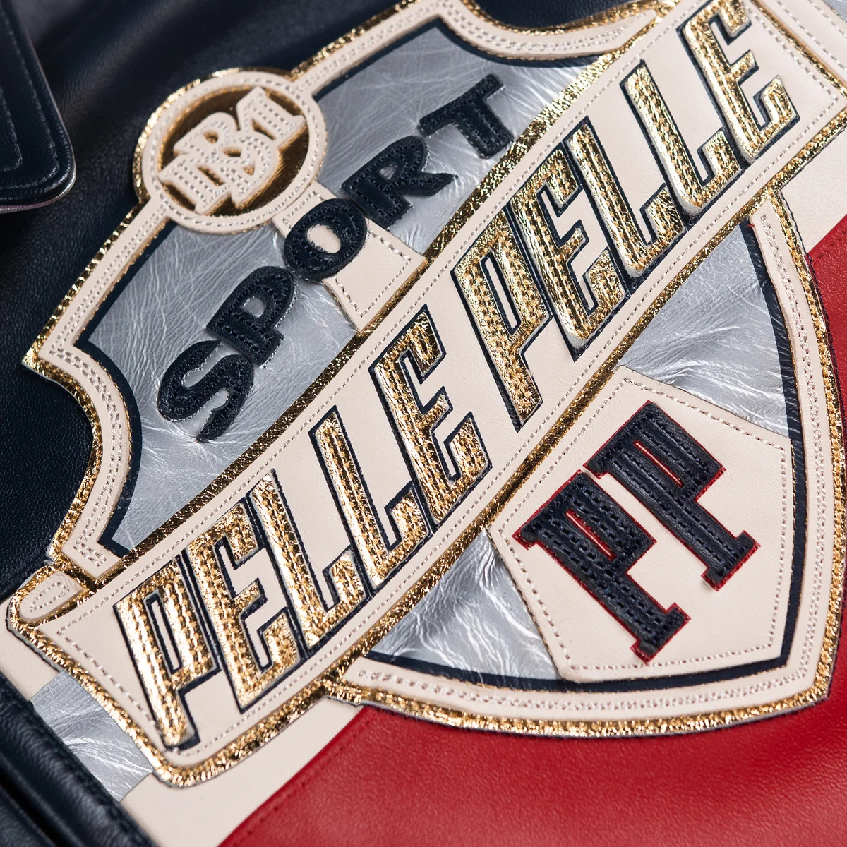 Close view of the sport jacket's shield chest patch with cream applique, metallic gold edging, silver panel and stitched black lettering.