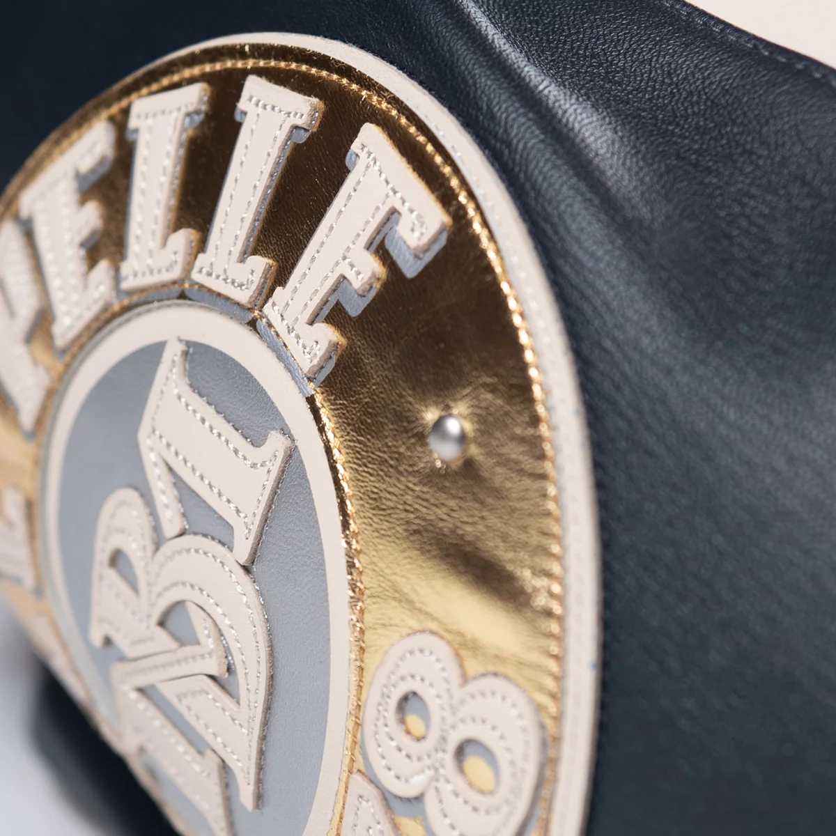 Detail of a circular sleeve patch on a black leather sport jacket, showing metallic gold panels, cream lettering and visible stitching.